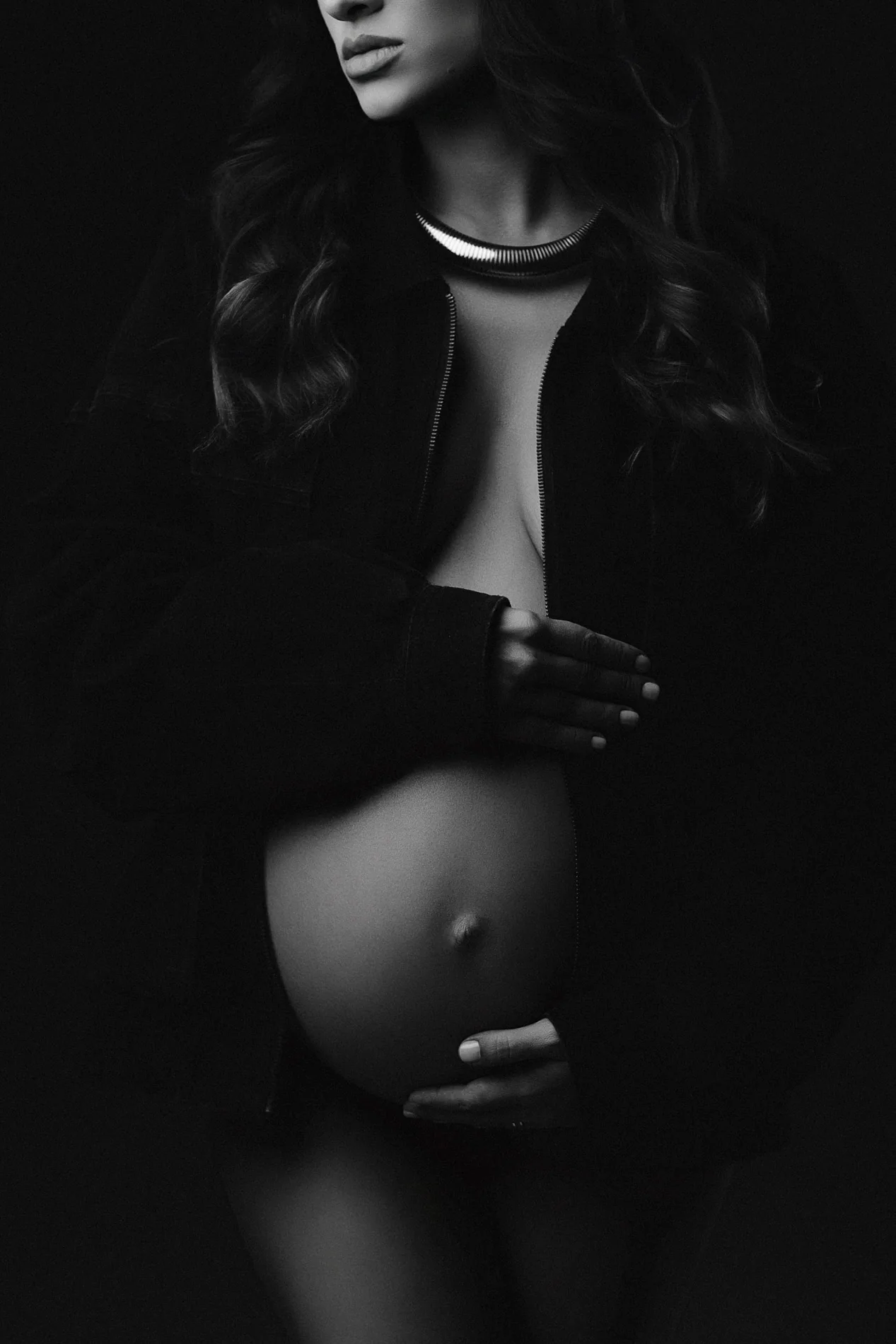 Retrato em preto e branco de gestante com roupa escura, mãos na barriga