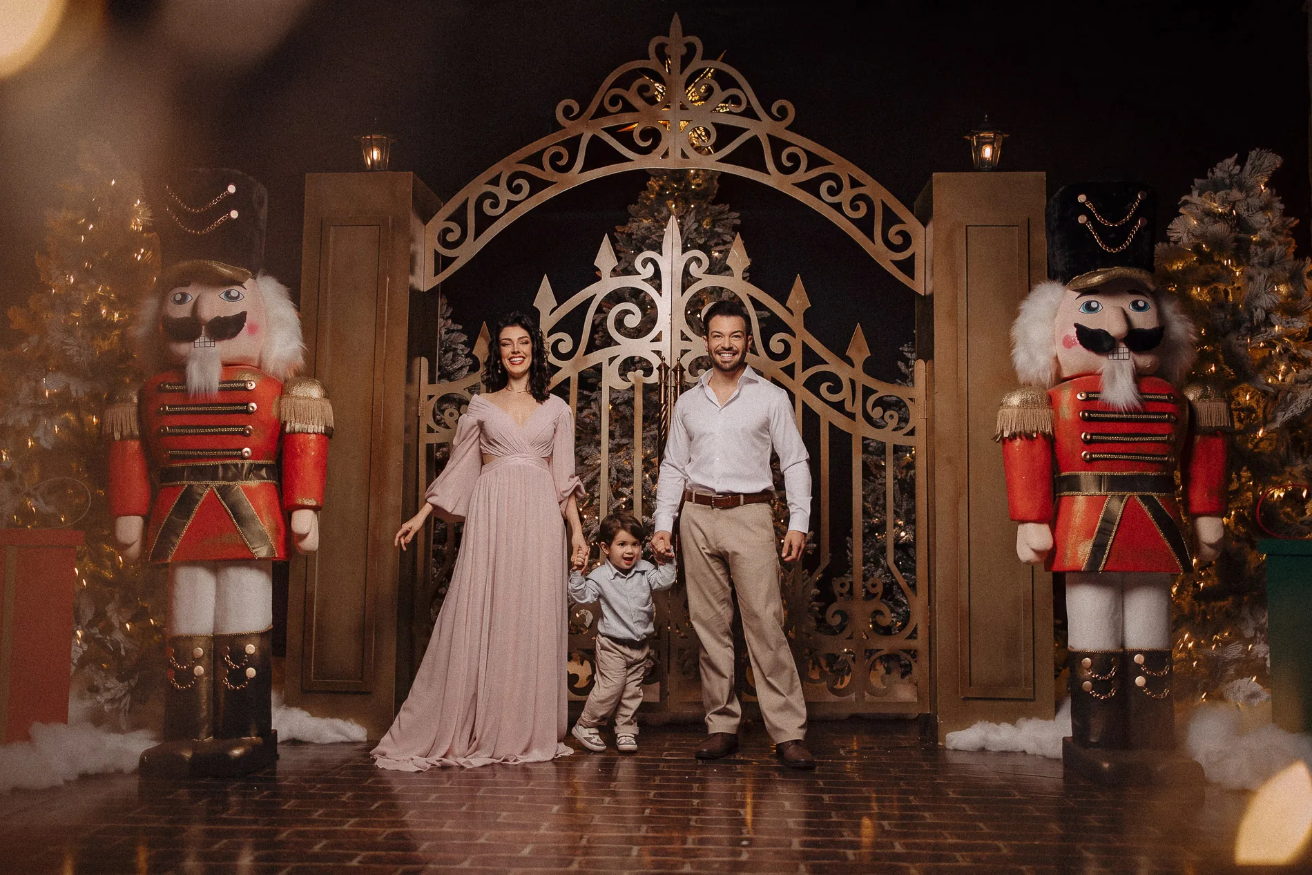 Família de mãos dadas diante do portão dourado e dos quebra-nozes do cenário de Natal.