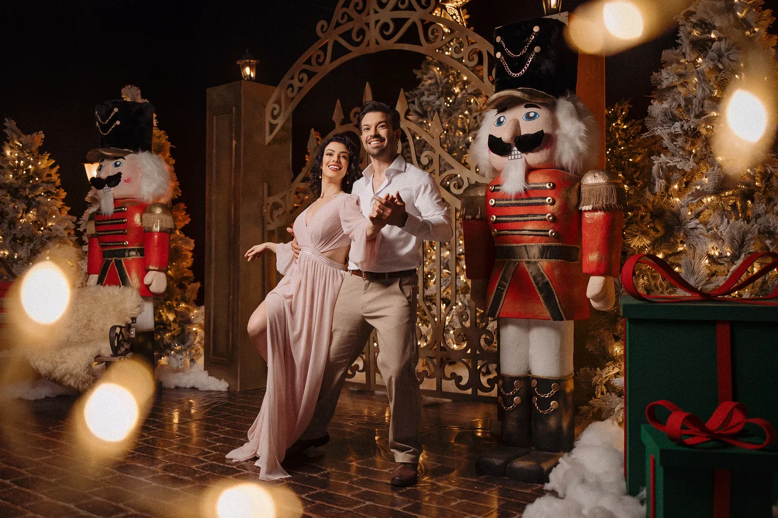 Casal no cenário do Especial de Natal 2026, entre o portão dourado e os quebra-nozes